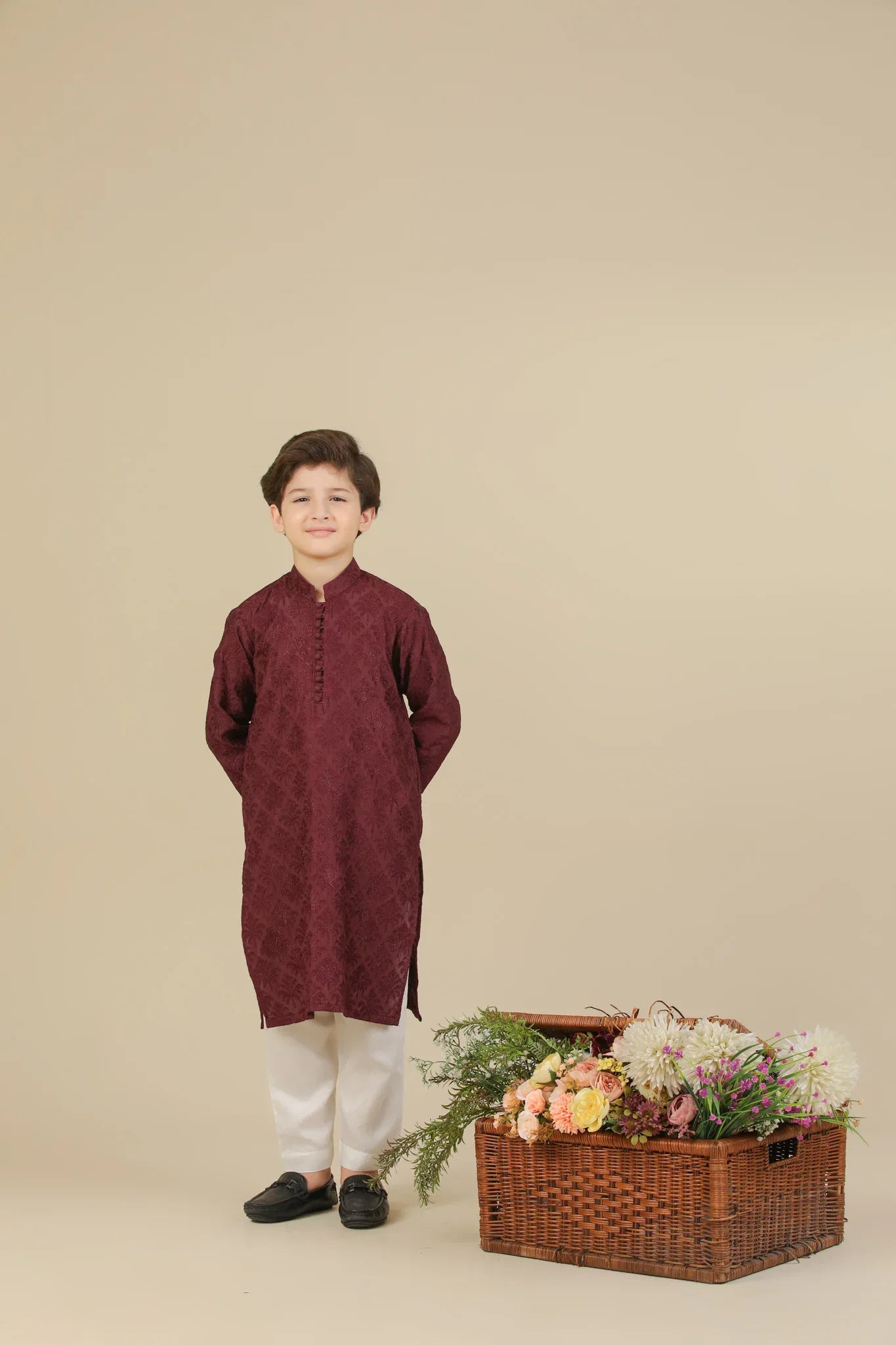 Child wearing a maroon raw silk embroidered kurta with white pants standing next to a basket of flowers on a beige background