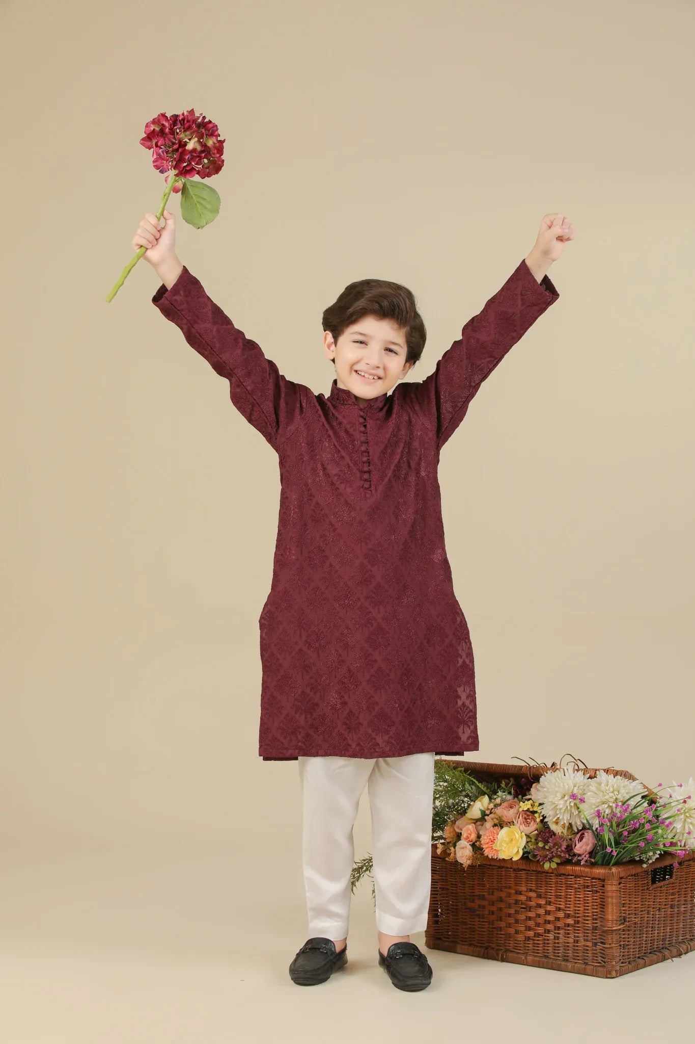 Child wearing a maroon raw silk embroidered kurta with white pants standing next to a basket of flowers on a beige background