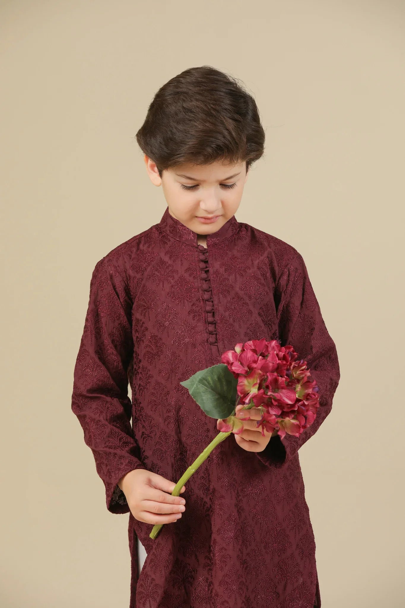 Child wearing a maroon raw silk embroidered kurta with white pants standing next to a basket of flowers on a beige background