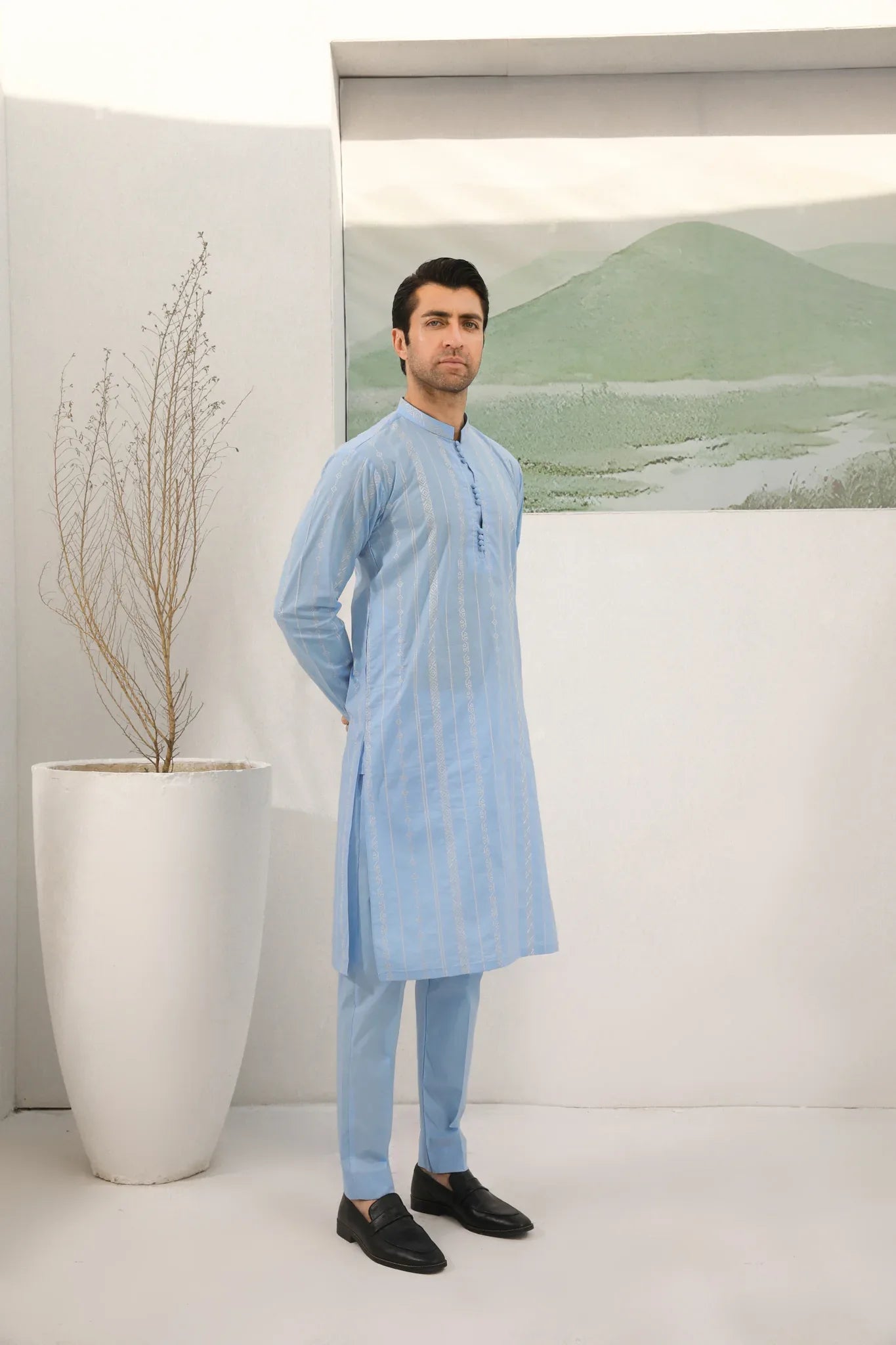 A man standing in a room wearing sky blue embroidered kurta and plain trouser in soft cotton fabric .