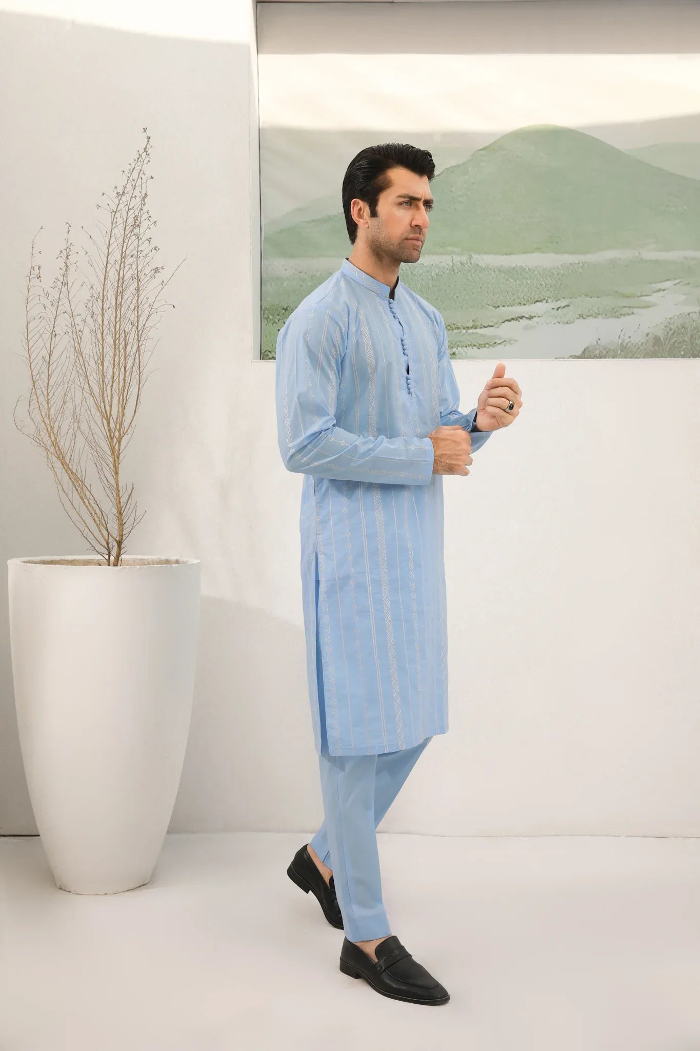 A man standing in a room wearing sky blue embroidered kurta and plain trouser in soft cotton fabric .