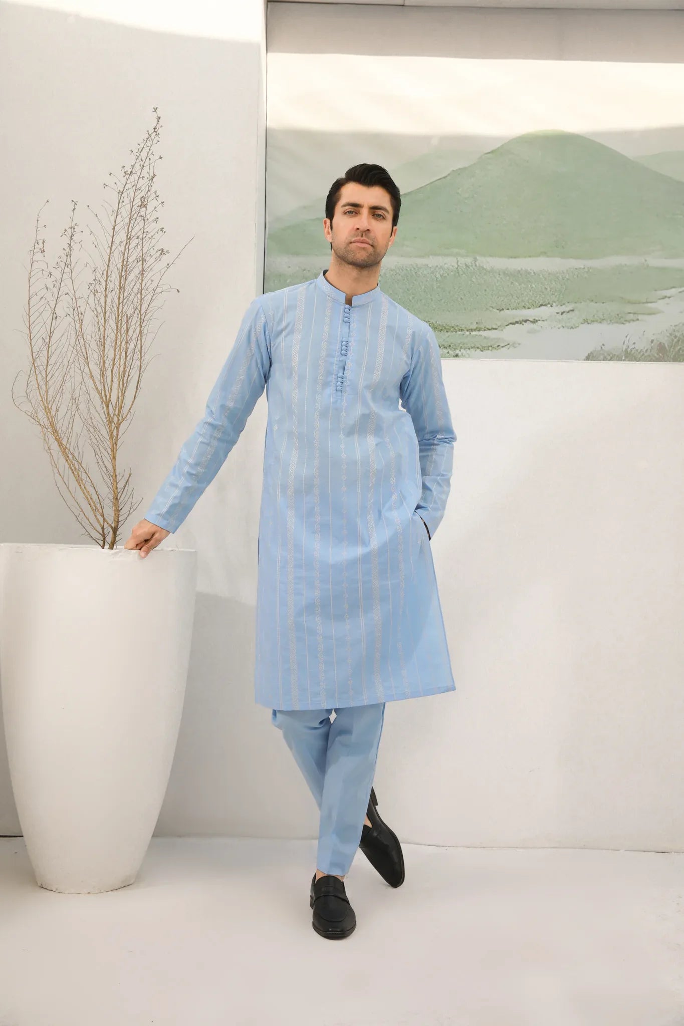 A man standing in a room wearing sky blue embroidered kurta and plain trouser in soft cotton fabric .