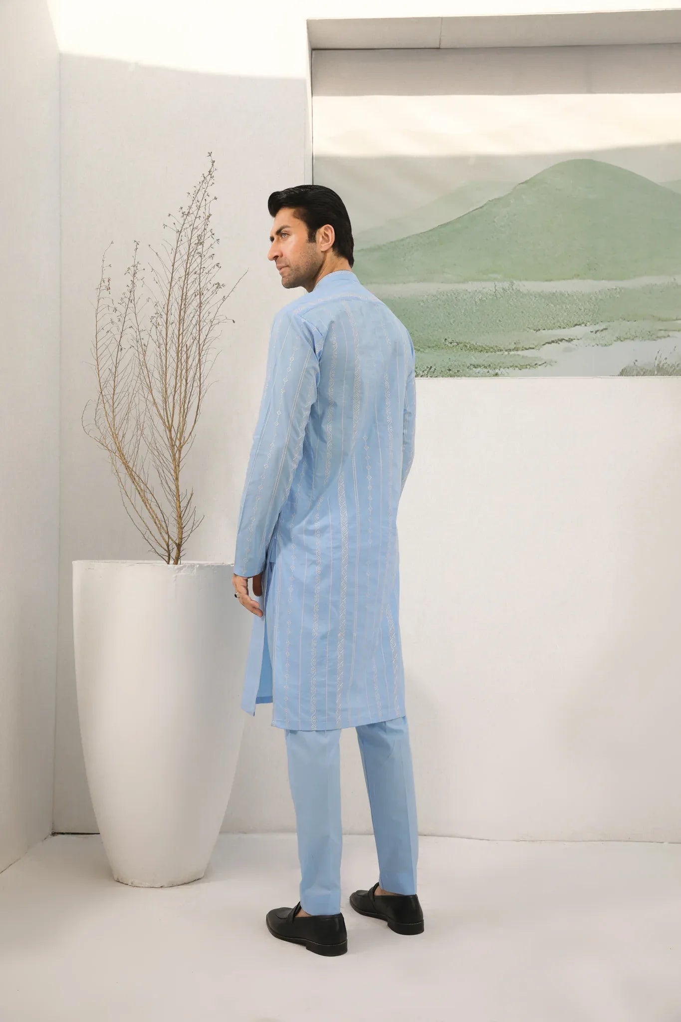 A man standing in a room wearing sky blue embroidered kurta and plain trouser in soft cotton fabric .