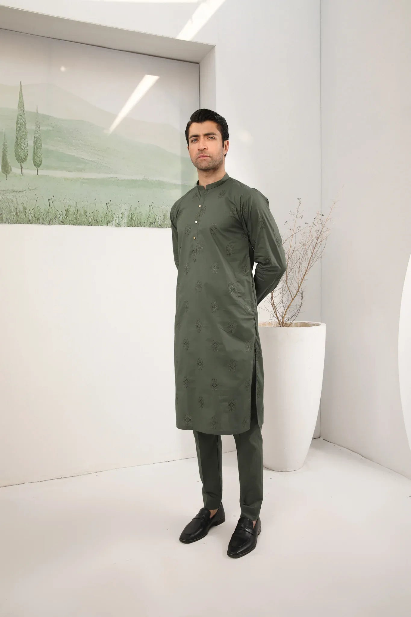 A man standing against a backdrop wearing a army green emnroidered kurta trouser.
