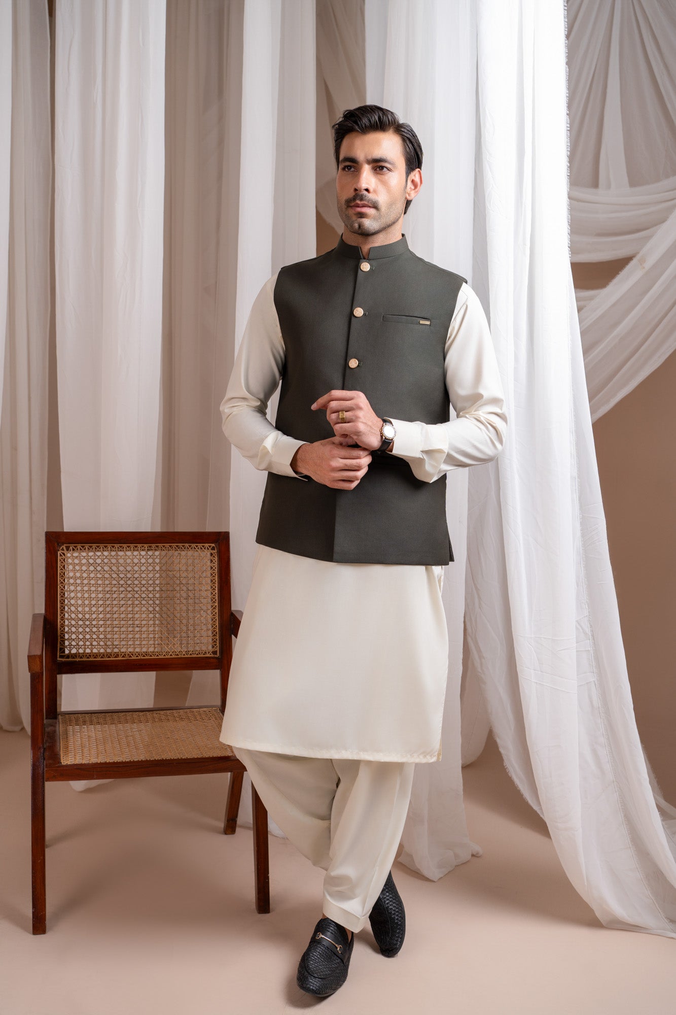 Man wearing a army green suiting waistcoat over bosky white kameez shalwar with a neutral background
