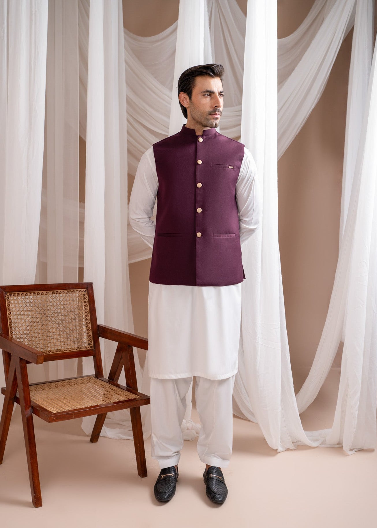 Man wearing a maroon waistcoat over a white kurta with white pants, standing in a room with curtains and a chair.