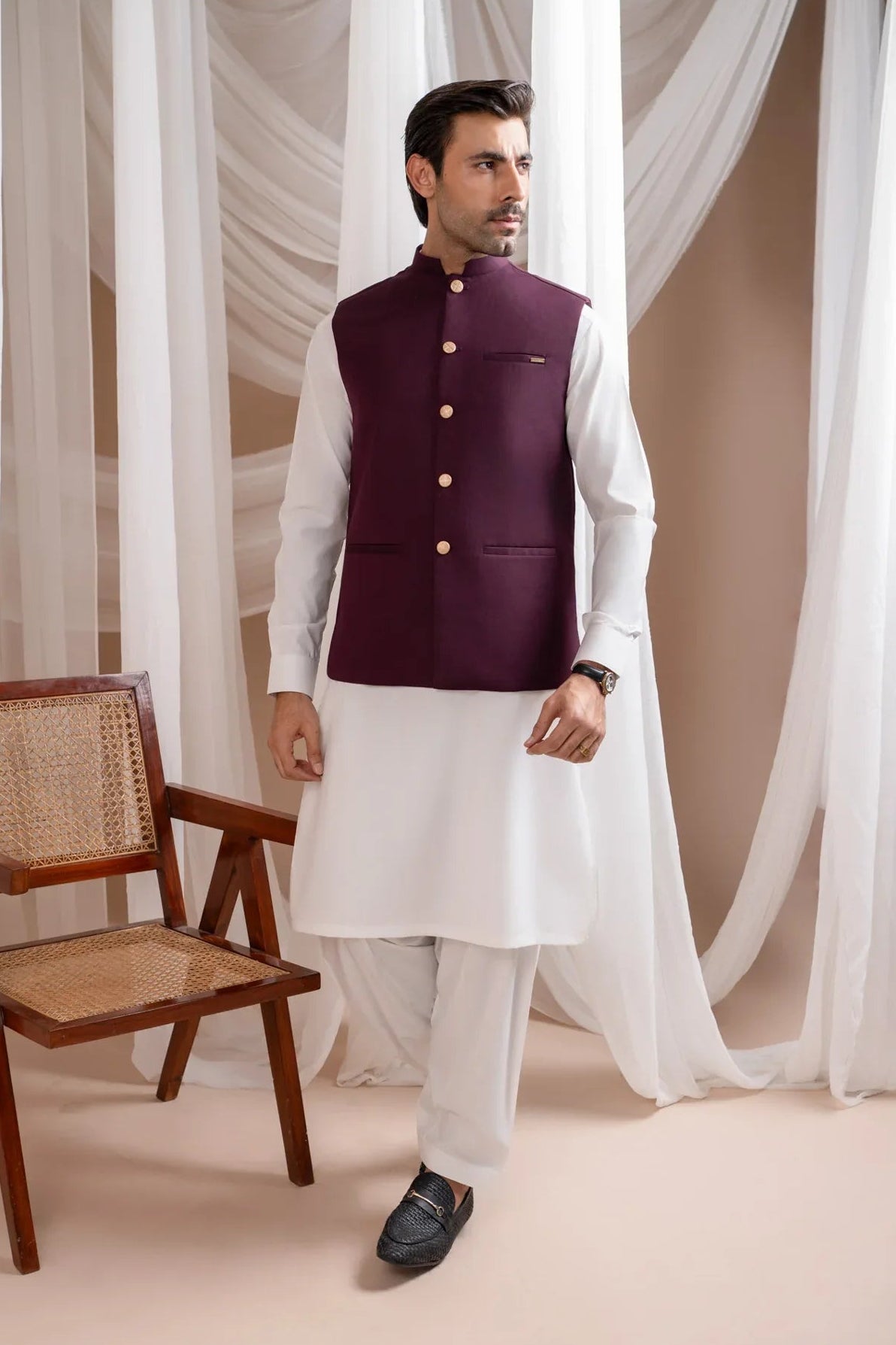 Man wearing a burgundy suitinf waistcoat over an off white kameez shalwar, standing in a room with curtains and a chair.