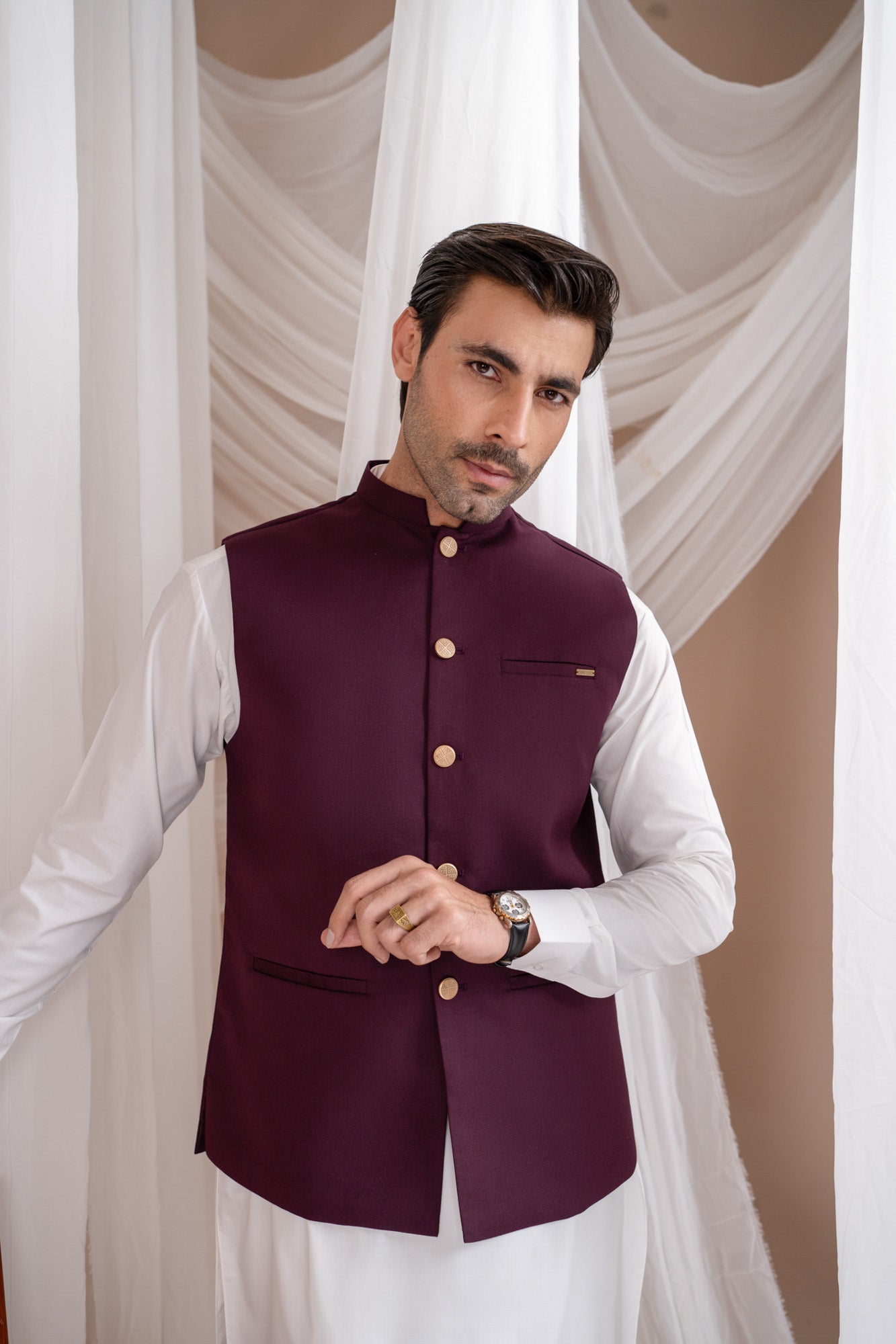 Man wearing a burgundy suitinf waistcoat over an off white kameez shalwar, standing in a room with curtains and a chair.